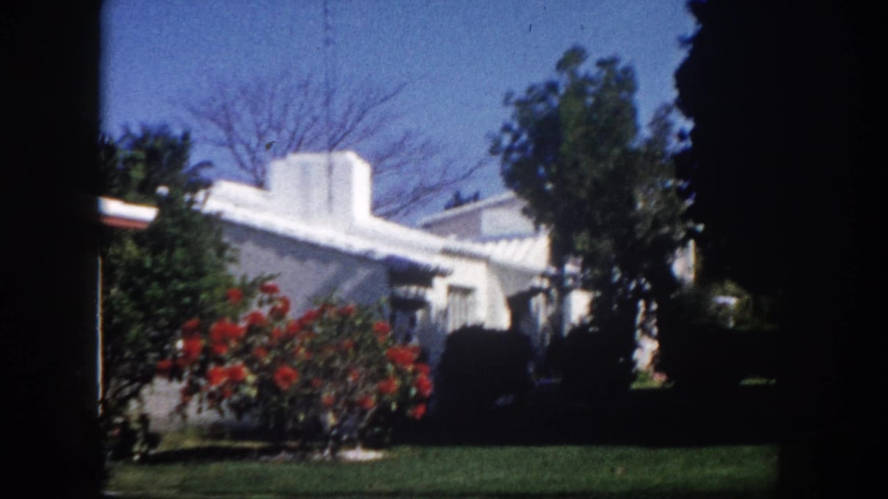 End frame showing a white flat-roofed stucco ranch house exterior with vivid red flowering shrub and green lawn under clear sky, Tampa Florida circa 1960.