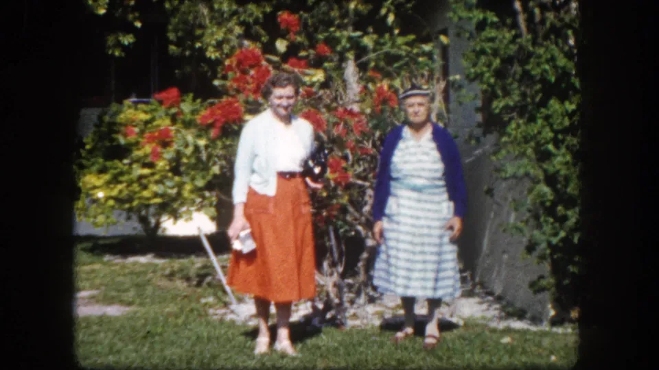 Two older women posing in a subtropical garden, Tampa Florida c. 1960: one in white top and orange midi-skirt, one in blue-striped dress with navy cardigan.