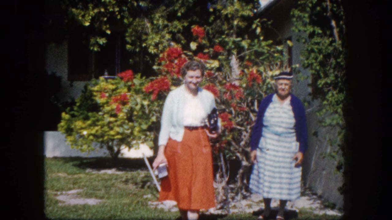 Two women stand on a sunny lawn before a red bougainvillea; one in an orange skirt and white top, the other in a blue-and-white striped dress with a navy cardigan.