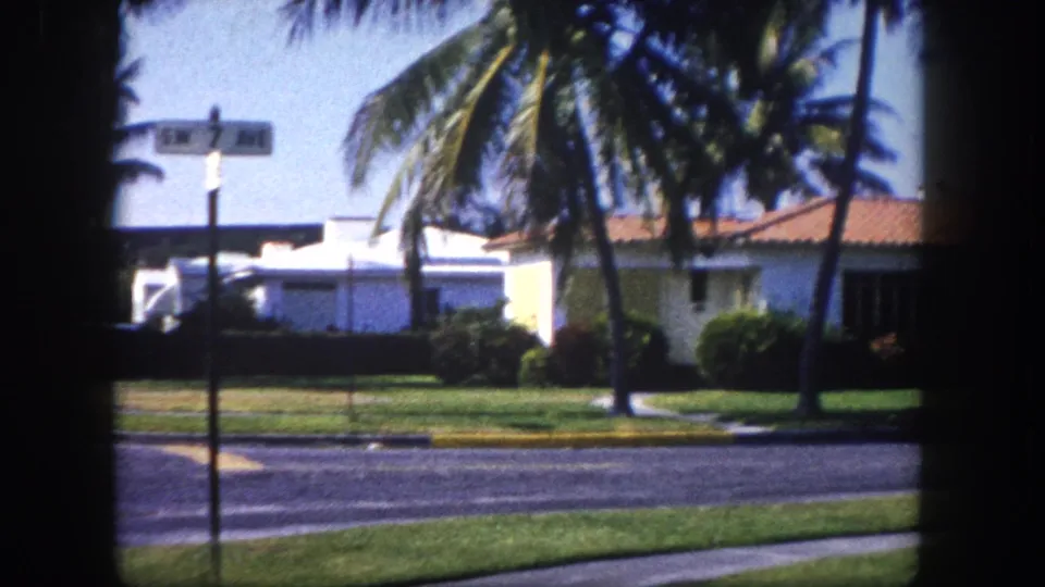 A palm-lined residential street in a Florida neighborhood, c. 1960, showing mid-century low-rise architecture under a clear blue sky, filmed on 8mm.