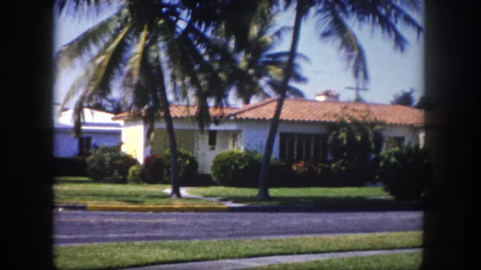 A mid-century white stucco house with terracotta tile roof and palm trees in a Florida neighborhood, c. 1960, filmed on 8mm color stock.