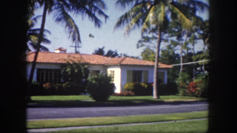 Continued pan of white stucco house with terracotta tile roof, palm trees, and flowering shrubs on a residential street in Tampa, Florida, circa 1960.