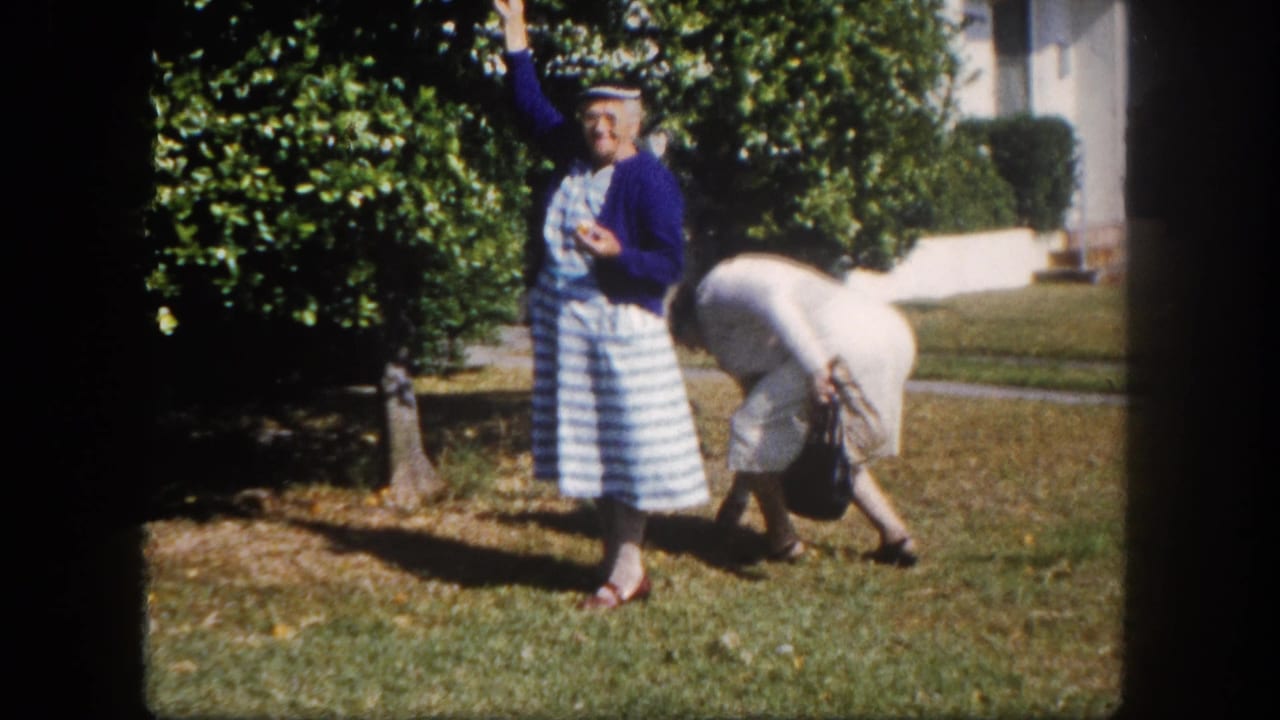 Two women outdoors; one in blue raises an arm in a wave while companion in white bends toward the grass, large green shrubs behind.