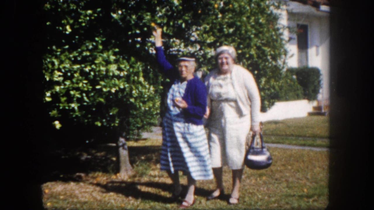 Two older women walking toward camera on a suburban lawn, Tampa, Florida, circa 1960; woman in blue cardigan waves while companion in white dress walks alongside.