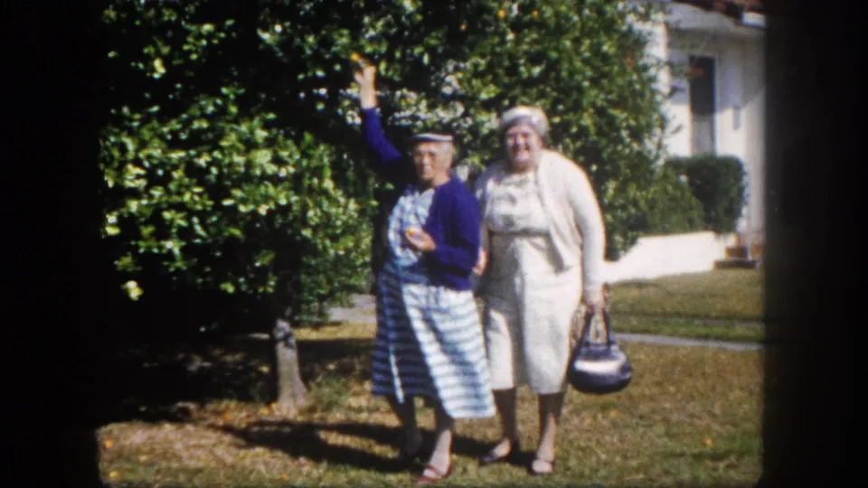 Two older women walking toward camera on a suburban lawn, Tampa, Florida, circa 1960; woman in blue cardigan waves while companion in white dress walks alongside.