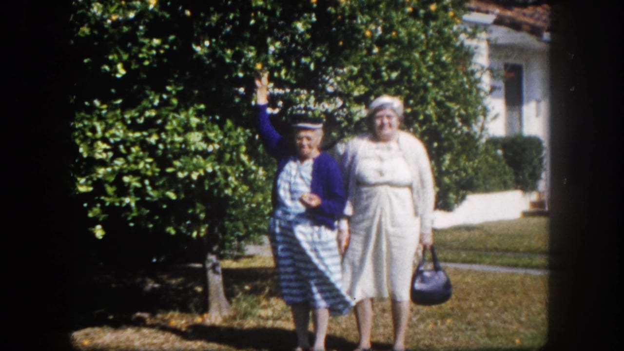 Two older women posing on a suburban lawn, Tampa, Florida, circa 1960; woman in blue cardigan waves while companion in white dress holds a dark handbag.