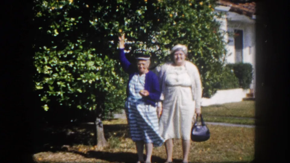 Two older women posing on a suburban lawn, Tampa, Florida, circa 1960; woman in blue cardigan waves while companion in white dress holds a dark handbag.