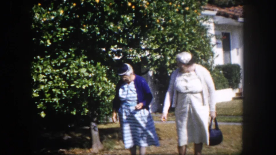 Two elderly women walking in a suburban garden in Tampa, Florida circa 1960; one in a blue cardigan and striped dress, the other in lighter clothing carrying a dark handbag.