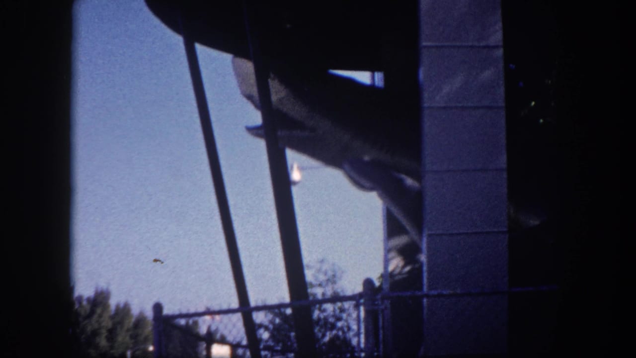 Rear section of a grey shark amusement ride figure on diagonal support arms, framed by a concrete-block pillar and chain-link fence at the 1960 Tampa State Fair.
