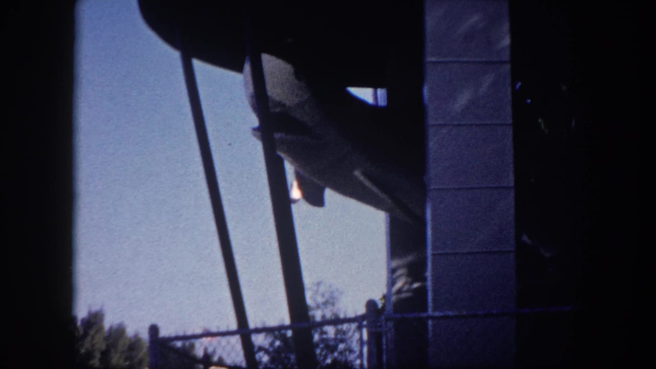 Shark fairground ride model seen from behind, suspended on diagonal support arms beside a central concrete pillar, with chain-link fence and trees in the background.