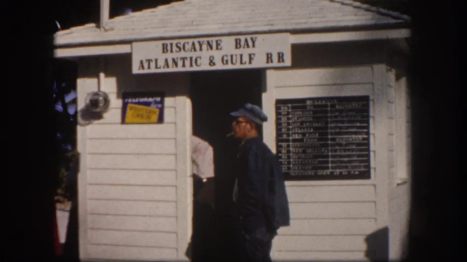 Man at the Biscayne Bay Atlantic & Gulf RR station building, Crandon Park area, Florida, circa 1960; schedule board and branded signage on white clapboard exterior.