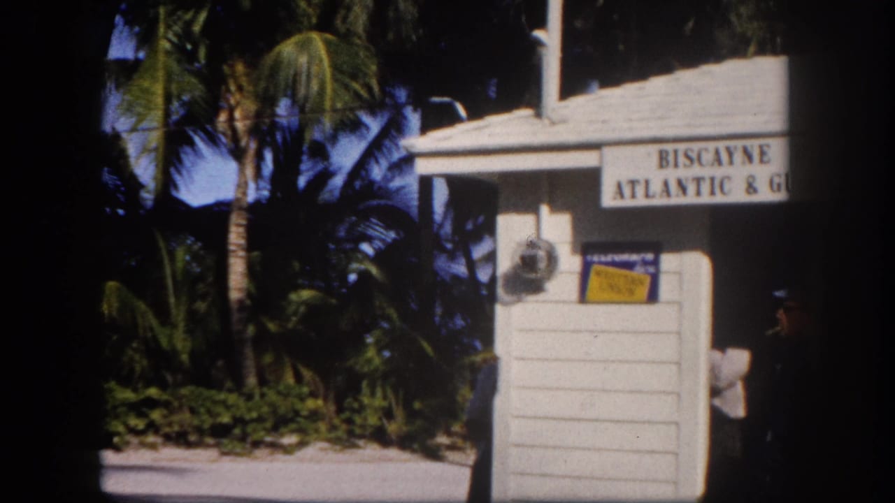 White wooden railroad station booth with partial Biscayne Atlantic & Gulf sign, dense palm trees behind, uniformed figure at right edge.