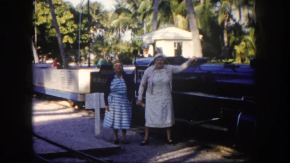 Two adult women in period dresses pose near a tourist tram in Florida, circa 1960, with palm trees and a white low-rise structure in the background