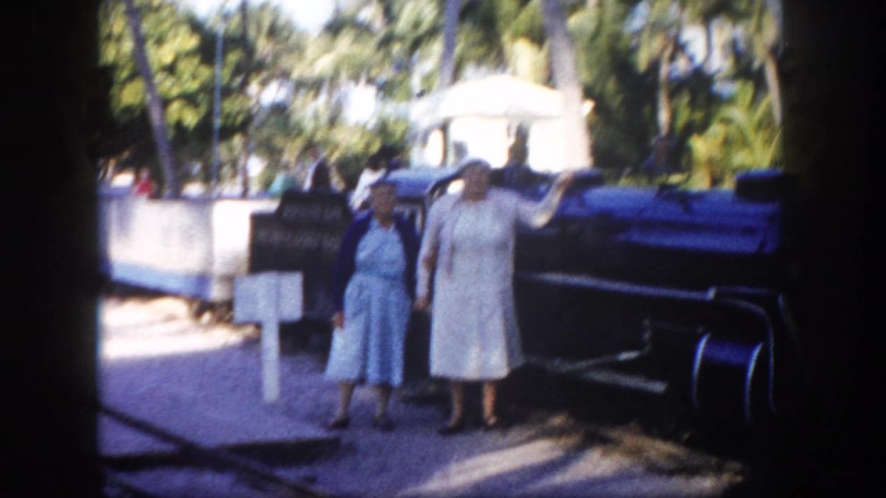 Two women pose beside a steam locomotive at what appears to be a Florida tourist attraction, circa 1960; palm trees and a white sign are visible behind them.