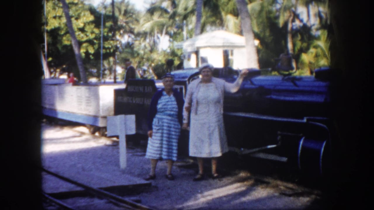 Two women beside a dark steam locomotive at a tropical location; one extends her arm toward the train.