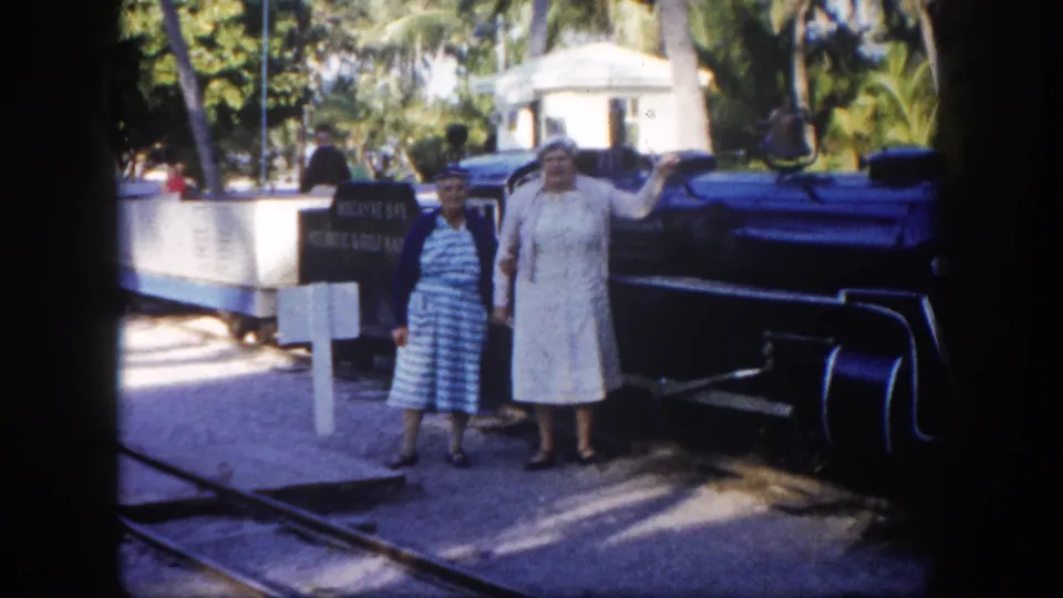 Two women pose together beside a steam locomotive at a tropical Florida tourist site, circa 1960; a white sign and palm trees are visible in frame.