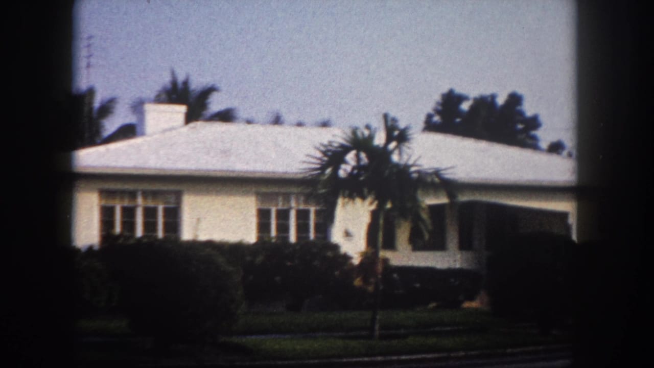 Pan continuing right: 1960s single-story Florida house with smaller palm in midground and right facade emerging, 8mm archival footage.