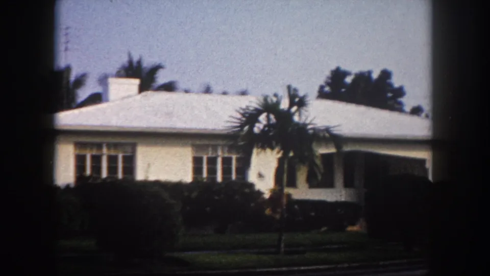 Pan continuing right: 1960s single-story Florida house with smaller palm in midground and right facade emerging, 8mm archival footage.
