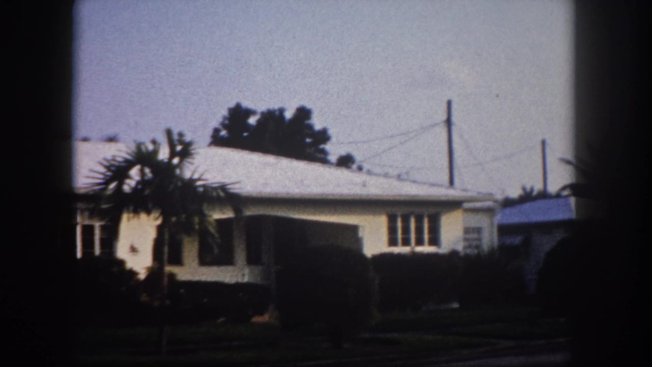 Pan end frame: right end of a 1960s Florida cream stucco house with utility poles, power lines, and a partial white vehicle at frame edge, 8mm archival.