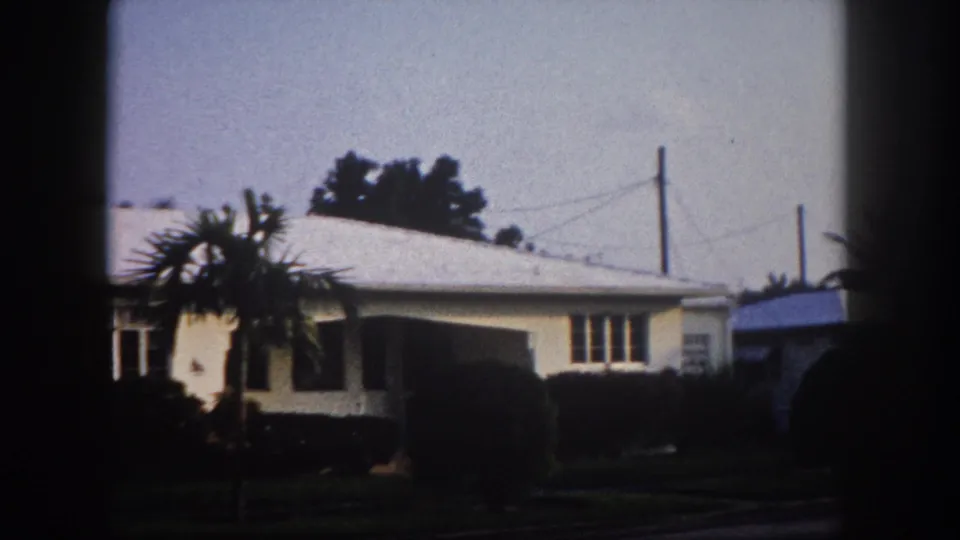 Pan end frame: right end of a 1960s Florida cream stucco house with utility poles, power lines, and a partial white vehicle at frame edge, 8mm archival.