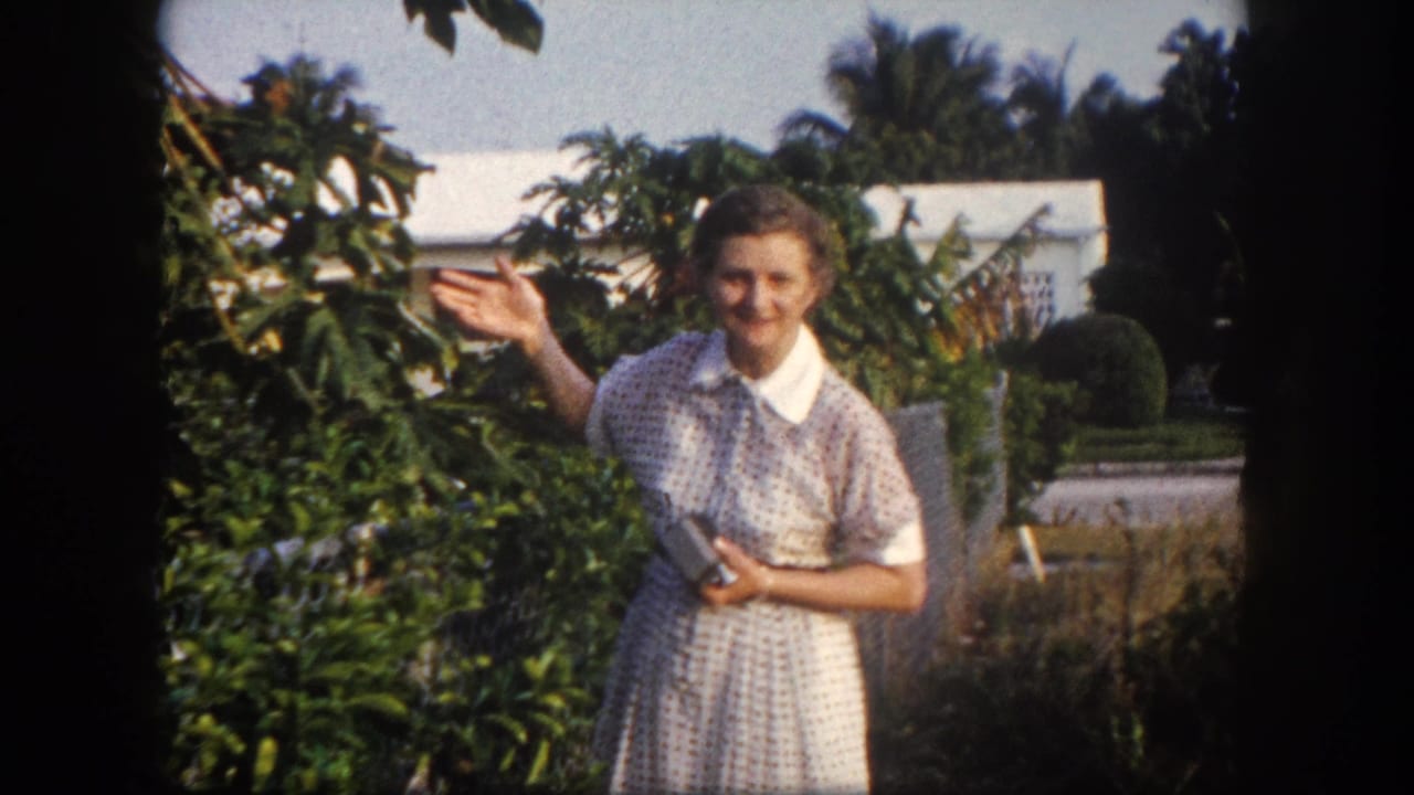 1960 Tampa home-movie frame of an adult woman smiling and raising one hand near leafy garden plants and a house.