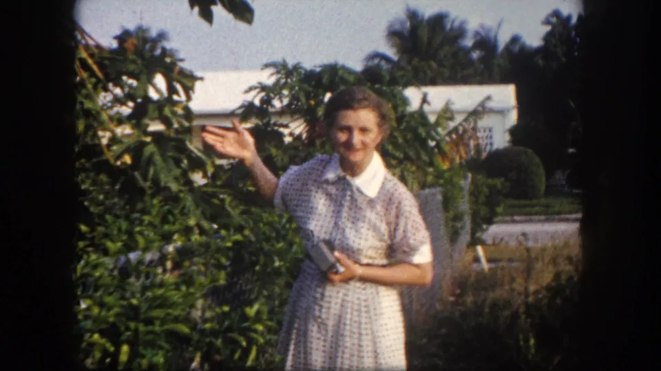1960 Tampa home-movie frame of an adult woman smiling and raising one hand near leafy garden plants and a house.