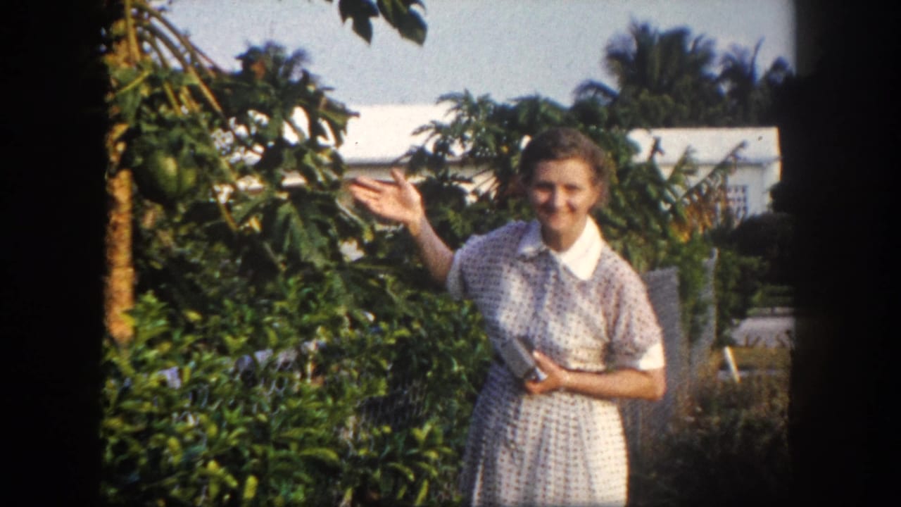 1960 Tampa home-movie frame showing an adult woman in a patterned dress standing among garden foliage with palms and a house behind.