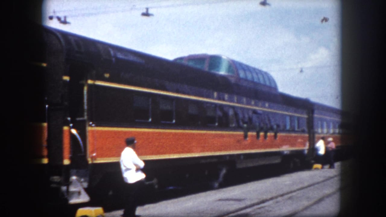 Person in white shirt and dark cap stands on station platform beside a stopped black and orange passenger train.