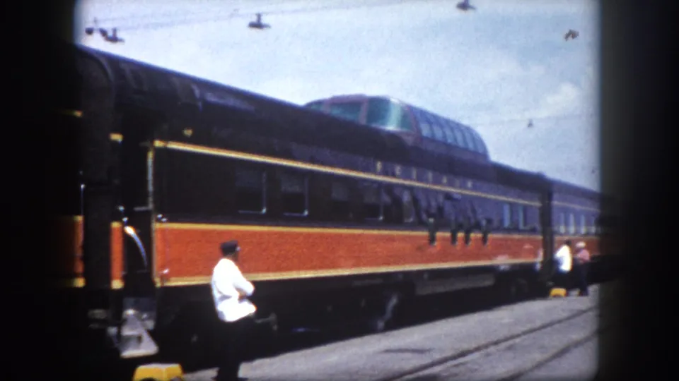 Home-movie frame showing a figure on the platform beside a mid-century passenger train with black-and-orange livery, Tampa, Florida, circa 1960.