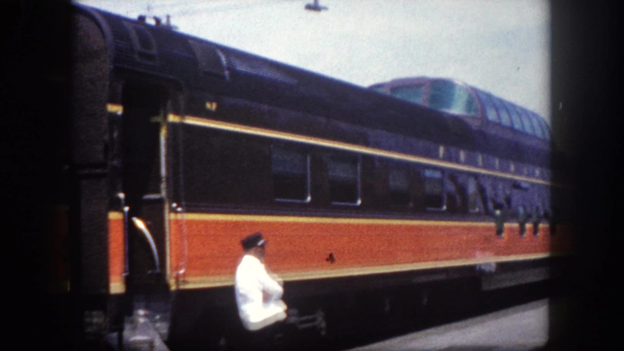 Figure in white shirt and dark cap stands on platform beside a stationary passenger train with orange-red and black livery.