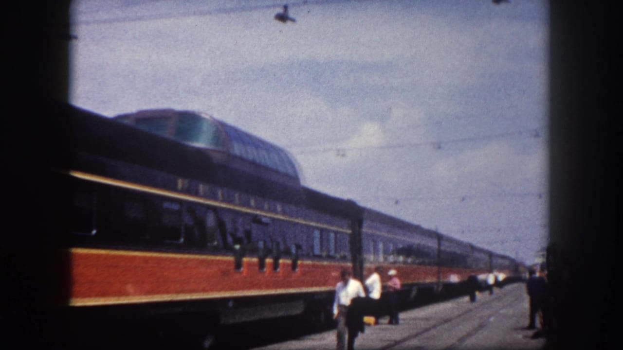 8mm home movie frame of a red-orange and dark blue passenger train at a Florida station platform, circa 1960, with platform figures in period attire and overhead catenary wires.