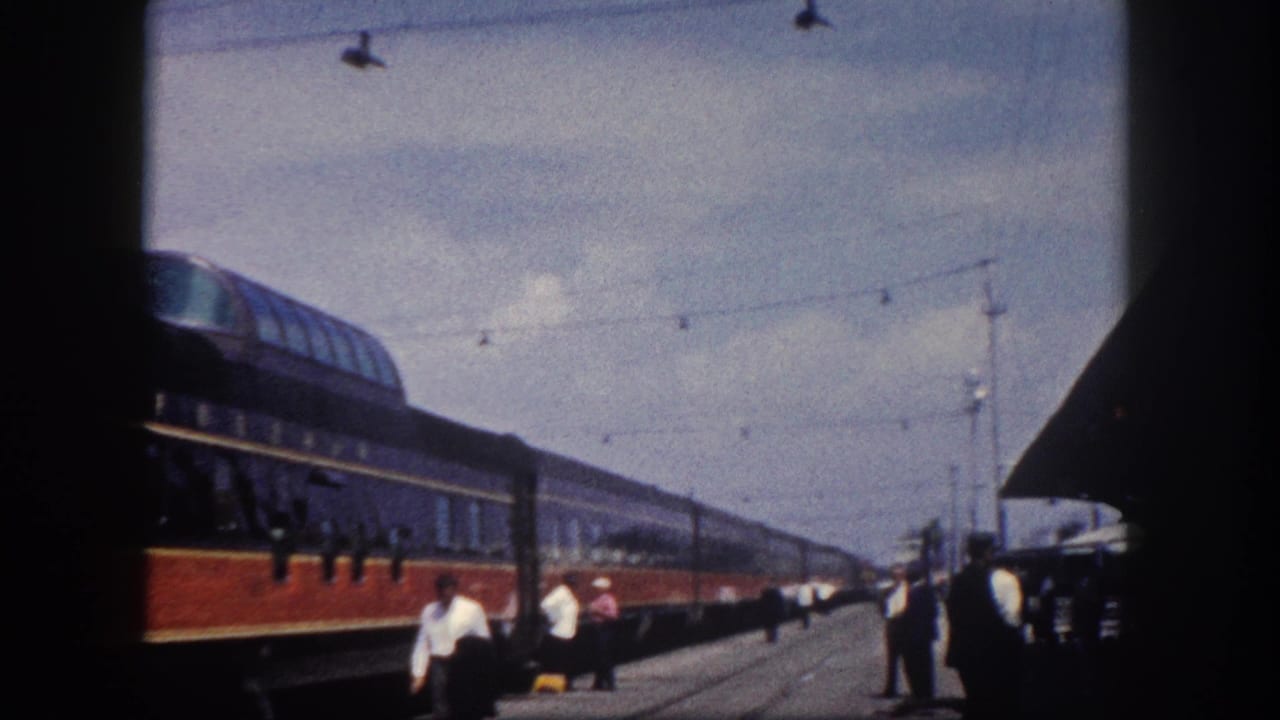 8mm frame of a long red-orange and dark blue streamliner at a Florida station, circa 1960; station structure, overhead wires, and platform figures visible at right.
