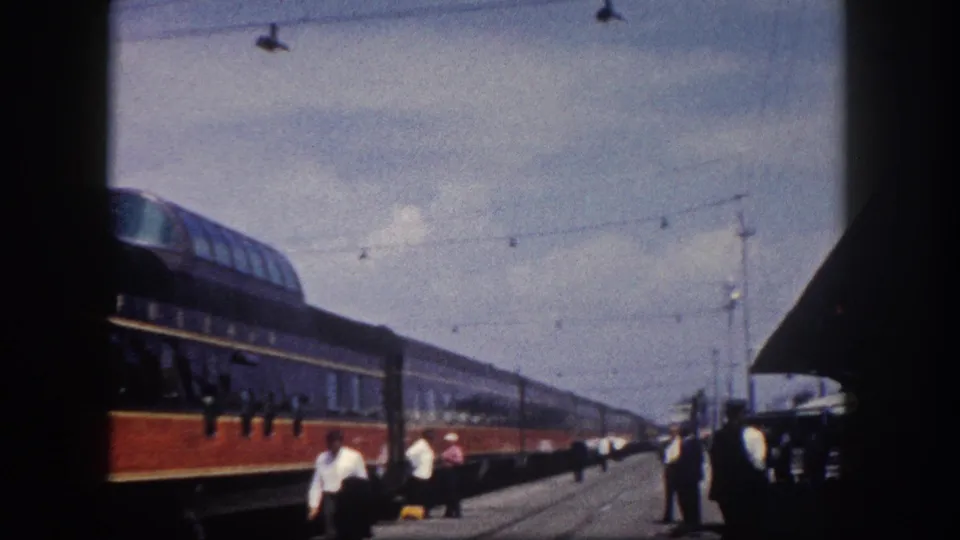 8mm frame of a long red-orange and dark blue streamliner at a Florida station, circa 1960; station structure, overhead wires, and platform figures visible at right.