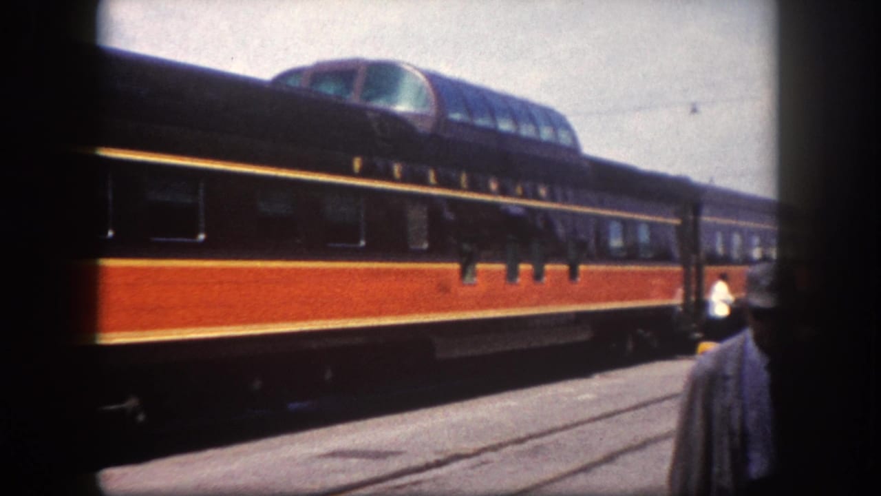 Orange and dark-liveried passenger train with dome observation car at a sunny station; a partially visible figure stands in the foreground.