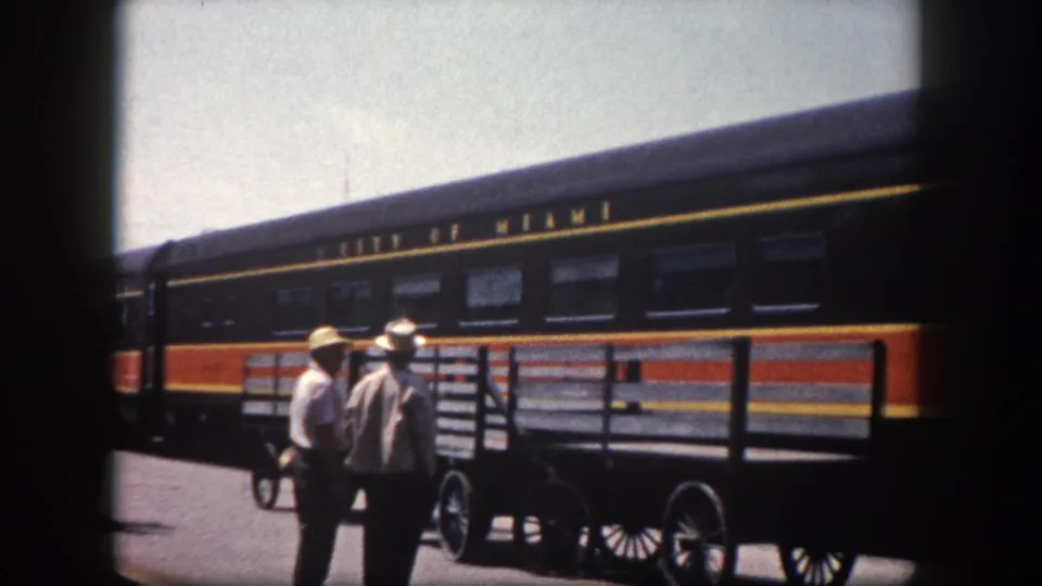 Two men in light shirts and wide-brimmed hats beside a spoked-wheel baggage cart, with the 'City of Miami' passenger railcar at a Tampa-area station, circa 1960.