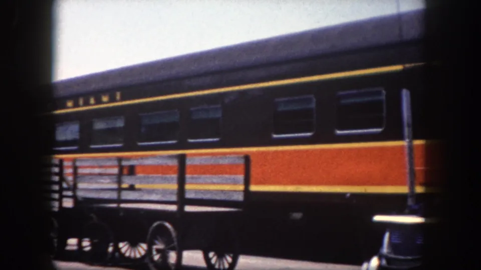 Detail of the 'City of Miami' railcar's distinctive orange, red and yellow livery with partial name lettering; a wheeled platform cart visible at lower left, Tampa area, circa 1960.