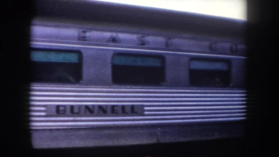 1960 Tampa area: stainless-steel railcar side with partial 'EAST COAST' lettering and 'BUNNELL' nameplate, a passenger dimly visible through window, shot on 8mm.