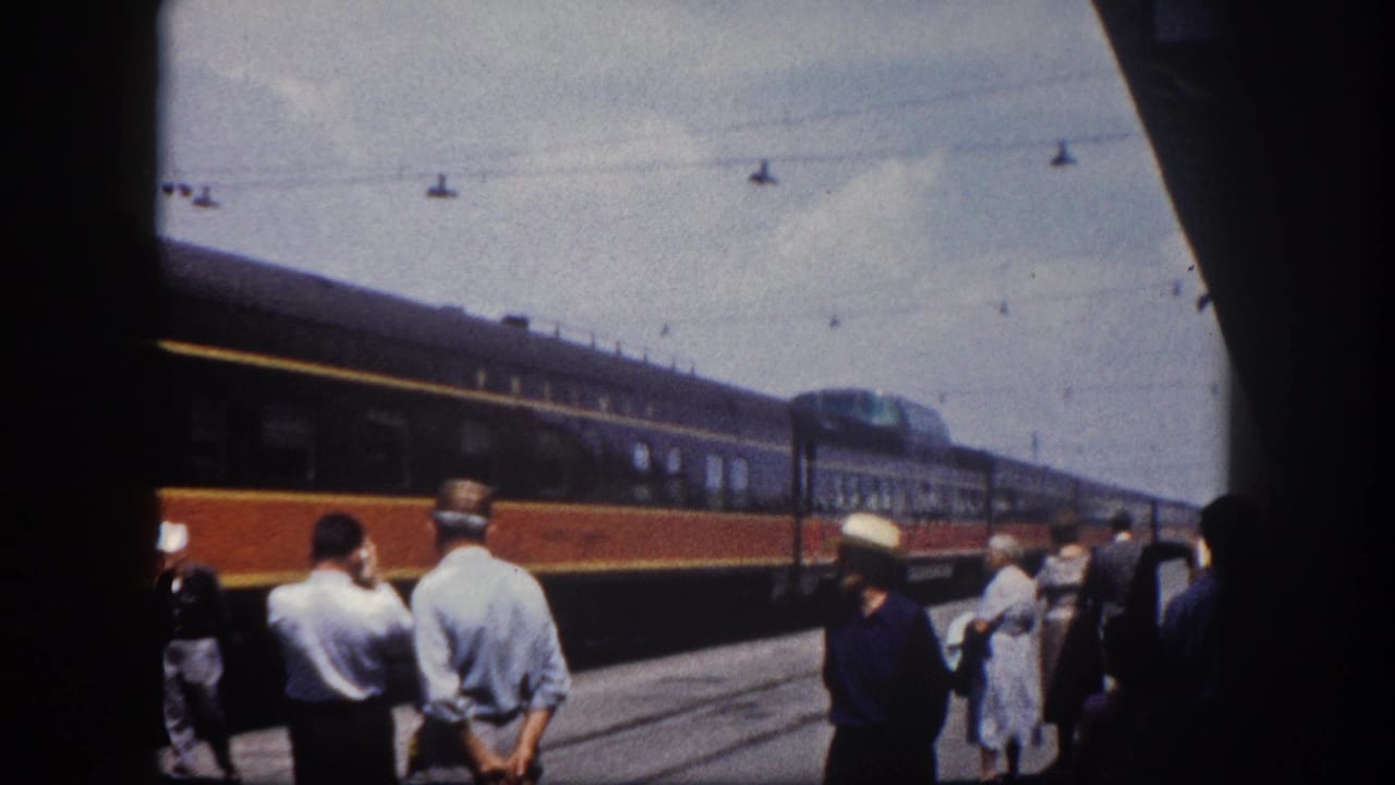 Passengers waiting on a Florida station platform, c.1960, as a two-tone passenger train passes; a woman in a dress is visible at the right edge.