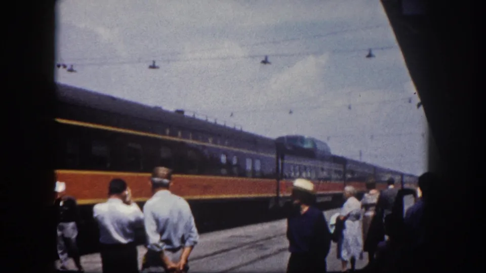 Passengers waiting on a Florida station platform, c.1960, as a two-tone passenger train passes; a woman in a dress is visible at the right edge.