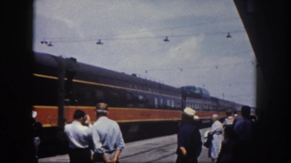 Men on a Florida station platform, c.1960, watch an orange-and-blue passenger train roll by; a brimmed straw hat is visible in the crowd.