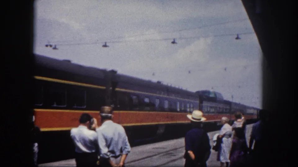 Florida station platform, c.1960: two men in light summer shirts and a woman in a dress watch an orange-and-blue passenger train pass.