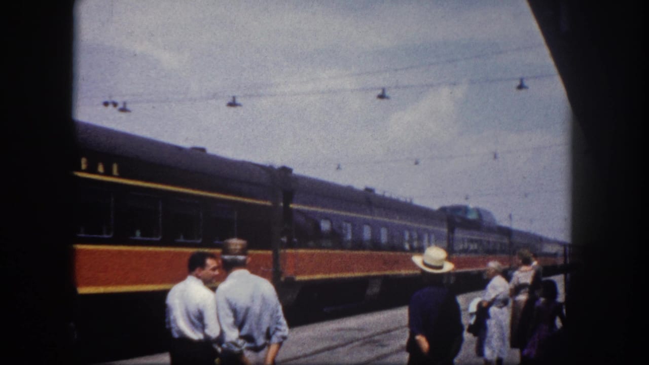 Passengers standing on a sunlit platform beside a dark red and gold-striped passenger train car