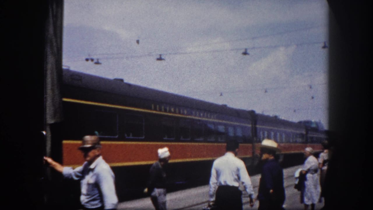 A uniformed railroad employee near an Illinois Central passenger car door, with boarding passengers in 1960s summer attire on the Tampa station platform
