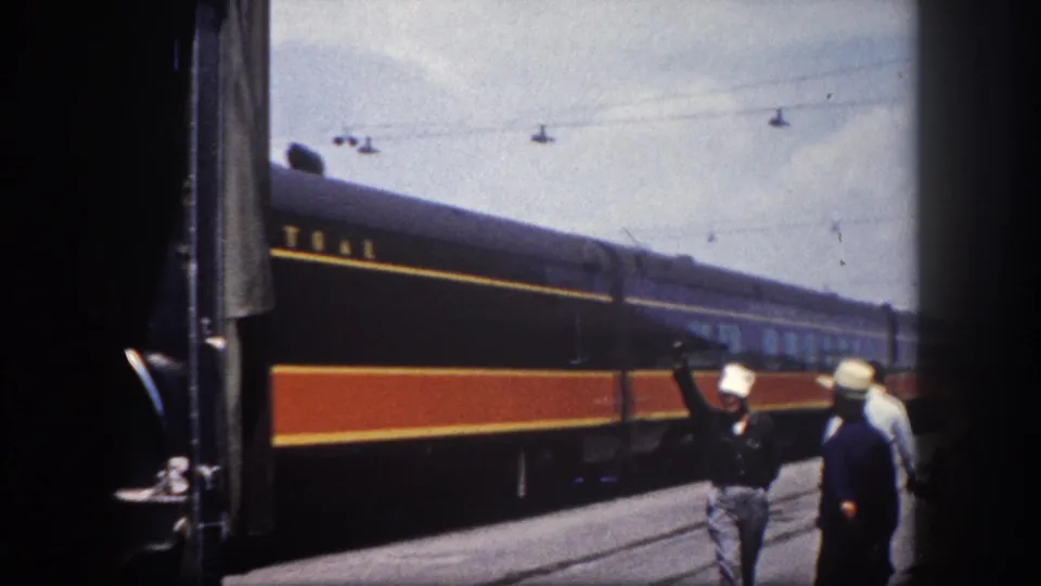 Two passengers in 1960s summer attire on a Tampa station platform as an Illinois Central train moves past, one wearing a white wide-brimmed hat