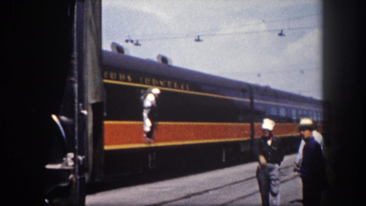 8mm home movie frame circa 1960, Tampa Florida: dark navy passenger car with orange and gold stripe rolls in as two men stand on the platform and a worker appears on the car steps.