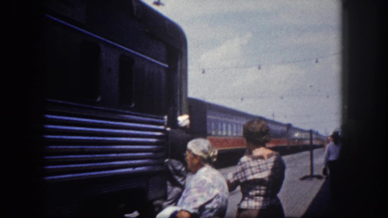 Two women on a train platform viewed from behind, a ribbed dark blue railcar filling the left side of the frame with an orange stripe visible.