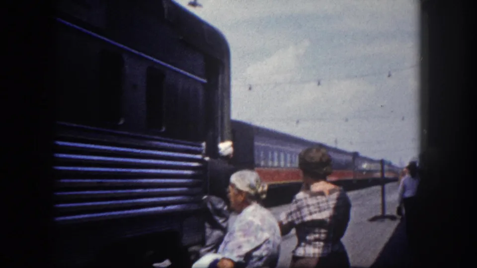 Close platform shot circa 1960, Tampa Florida: elderly woman in floral dress and companion in plaid stand beside the dark blue passenger car as it arrives.