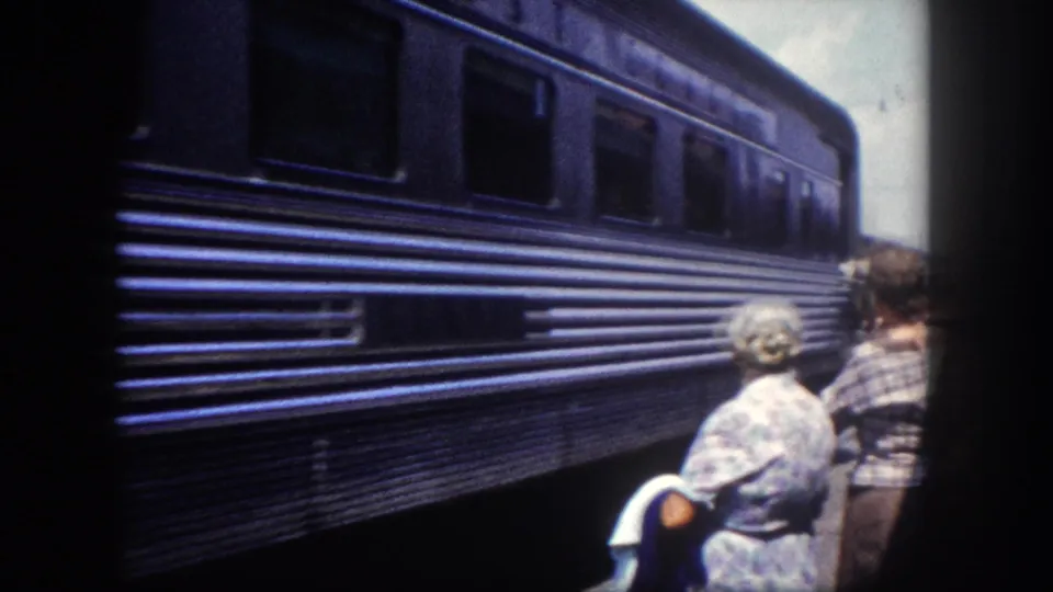 Final 8mm frame circa 1960, Tampa Florida: elderly woman in floral dress and companion in plaid stand close to the towering dark blue railcar, a third figure partially visible at frame edge.