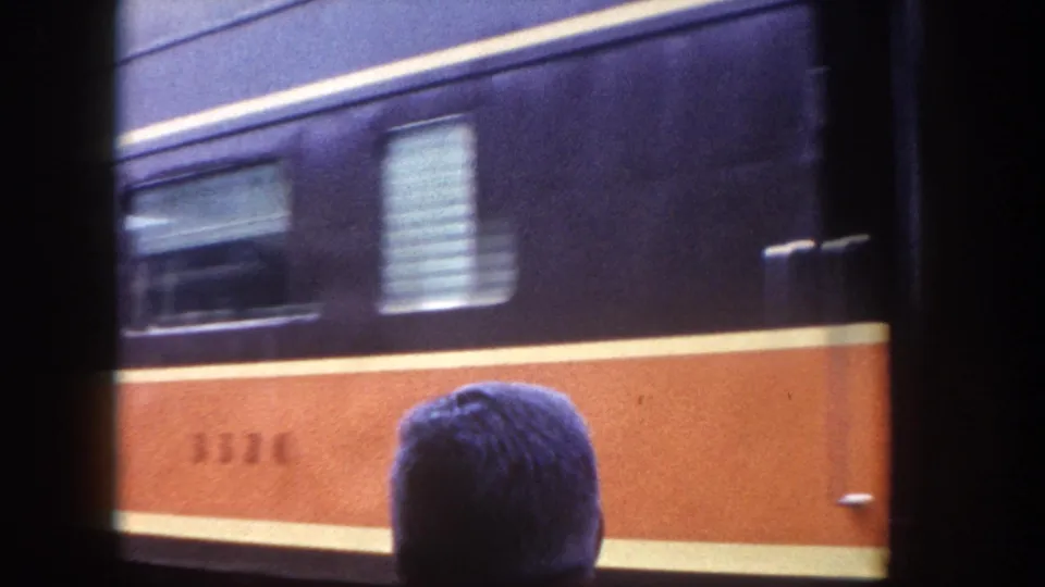 8mm home movie frame of a moving passenger railcar showing a car door and ventilation louvers, with a partial car number on the orange band and a bystander below, circa 1960.