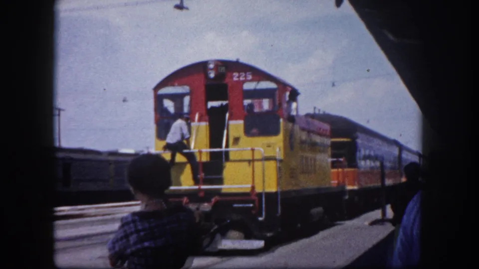 Platform observer in plaid top watches red and yellow locomotive 225 at Tampa station, circa 1960; overhead wires and additional orange rail cars visible in background.
